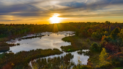 sunset on a beaver pone in NY  aerial perspective showing setting sun with clouds fall colors in the trees.  Reflections on the pond with tall grasses 