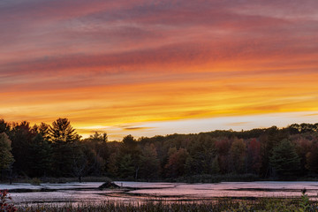 sunset on pond with beaver hut in the distance orange red yellow cloud streaks