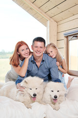 Young Caucasian family couple with baby daughter in a small modern rustic house with a large window. Lying on the bed, hugging, playing and looking out the window. Two Samoyeds. Weekend vacation.