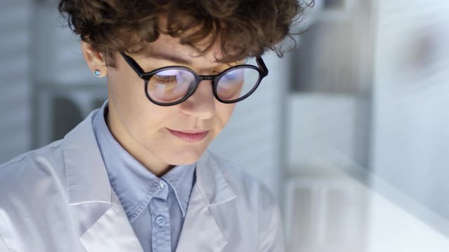 Close up shot of female doctor in lab coat and eyeglasses looking at transparent monitor while working on futuristic computer in office