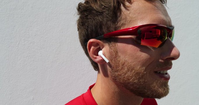 Young man putting on wireless earphones going running wearing sport sunglasses for fitness run outdoors. Happy active person wearing earbuds for exercise. Man listening to audiobook or podcast.