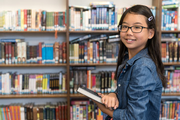 Back to school concept with a teenage Asian girl with eyeglasses standing with books in the school library