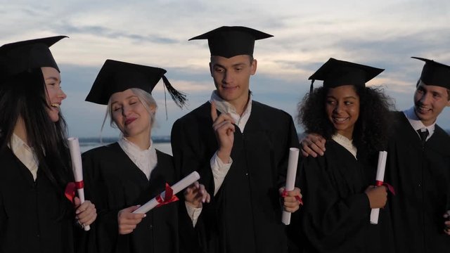 Mixed races happy students in classic graduation mantles and academic hats walking and talking.
