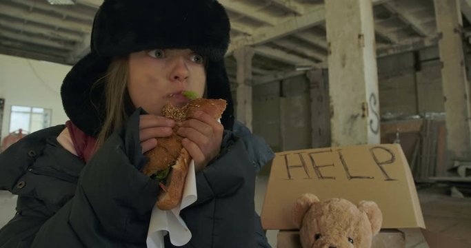 Portrait of a Caucasian homeless girl with dirty face and grey eyes greedily eating the bun. Hungry child in hat with earflaps living on the streets.