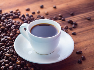 Composition of white coffee cup with black espresso and coffee beans pilled on brown table, close up view. Texture of wooden desk. Selective soft focus. Blurred background