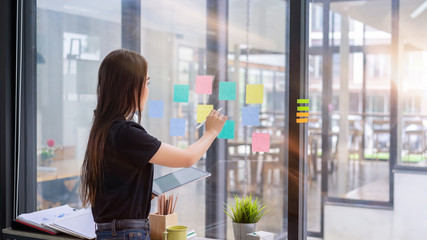 Creative businesswoman using tablet and putting her ideas on a sticky notes on glass wall at office.Business people meeting sharing idea concept.
