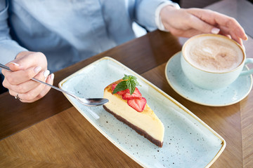 A young woman drinks coffee cappuccino in a restaurant and eats dessert. Piece of delicious cheesecake with strawberry and mint leaves on white plate. Restaurant menu