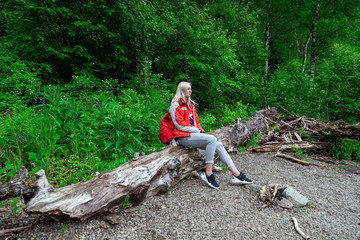 Beautiful blonde girl in a autumn jacket with a red backpack sits on a fallen tree in a forest or...
