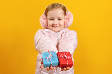 Happy little girl holds out small boxes with gifts. A child in a jacket and warm ear muffs on a yellow background. Christmas and new year concept