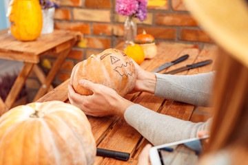 Halloween Preparaton Concept. Young couple sitting at table at porch making jack-o'-lantern man holding pumpkin with face drawn while woman holding smartphone to take photo