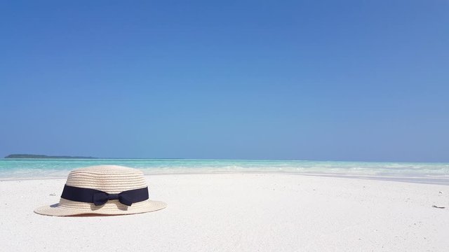 Low angle shot of beige tourist hat with black ribbon stipe around it. Hat laying on white sand beach and sea tides and blue clear sky on background.