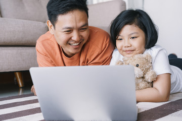 Asian father and daughter  using a laptop while lying on the carpet in the living room at home. The cute little girl hugs the doll and smile happily with his father.