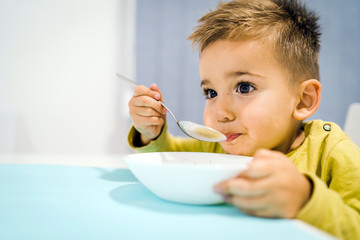Portrait of small boy child eating soup meal or breakfast having lunch by the table at home with spoon white kindergarten