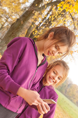 Outdoors leisure. One sister playing game on smartphone happy while another looking at screen cheerful standing in the park close-up