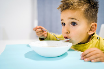 Portrait of small boy child eating soup meal or breakfast having lunch by the table at home with spoon white kindergarten
