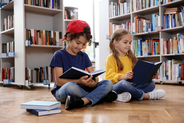 Cute little children reading books on floor in library - obrazy, fototapety, plakaty