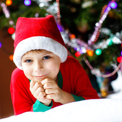 Little kid boy in santa hat with Christmas tree and lights on background. With colorful illumination and garland. Happy preschool child celebrating xmas, family holiday. Boy in nightwear.