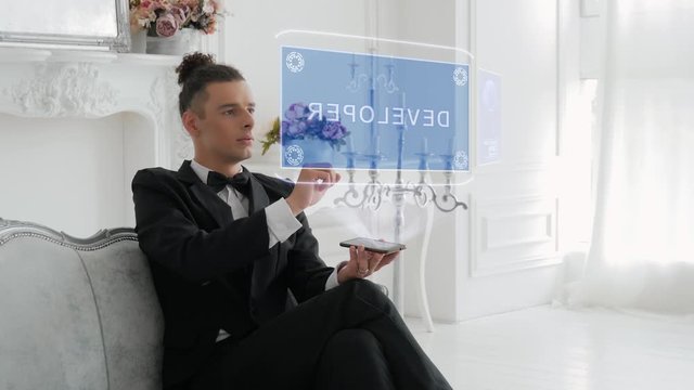 Guy businessman activates conceptual HUD hologram on smartphone with text Developer. Young man in a white shirt and a jacket with a holographic screen on a background of the luxury room
