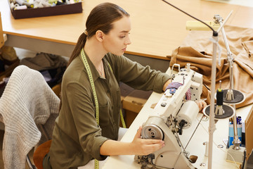 Serious young woman sitting and sewing using sewing machine in atelier