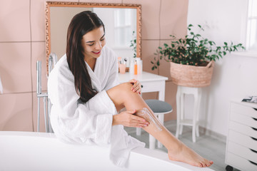 Beautiful young woman applying cream on her legs in bathroom