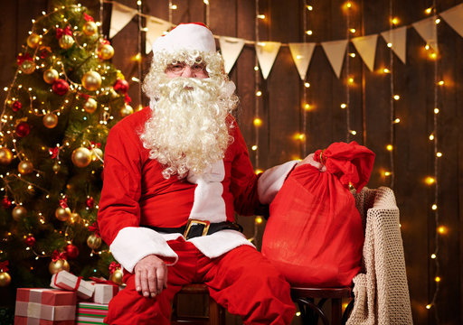 Santa Claus posing with bag of gifts, sitting indoor near decorated xmas tree with lights - Merry Christmas and Happy Holidays!