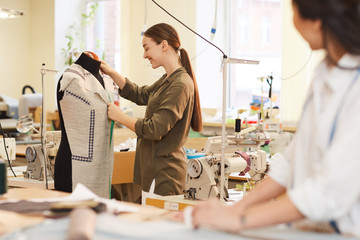 Young smiling designer trying on the jacket on mannequin and examining it before sewing she working...