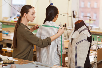 Young tailor measuring length of shoulders with tape measure of her customer while she standing...