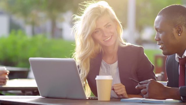 Tracking medium shot of attractive blonde young businesswoman talking to black male colleague and using laptop computer in outdoor cafe
