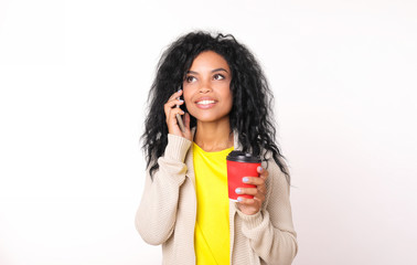 Busy as a bee. Busy African American woman in beige sweater is talking on the phone while holding a red paper coffee cup in her left hand, looking at the upper left corner and smiling.