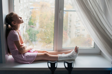 A teenage girl with a broken leg in a cast is sitting on a windowsill with crutches.