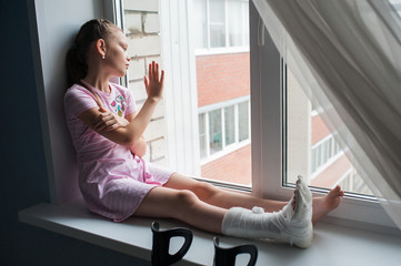 A teenage girl with a broken leg in a cast is sitting on a windowsill with crutches.