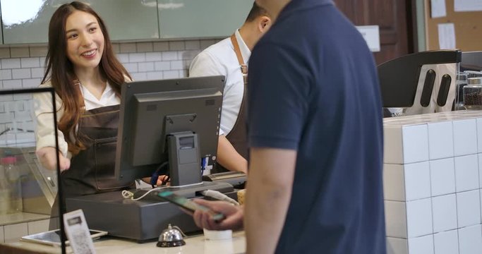 Beautiful asian woman serving a coffee to go to customer who uses smartphone to pay in coffee shop. QR code cashless payment concept.