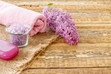 Obraz na płótnie Canvas Bottle with oil, bowl with sea salt, soap and towel with lilac flowers on wooden background.