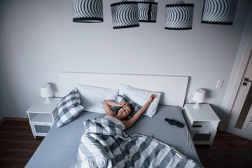 Stylish room. Woman with blindfold mask for a sleep is lying on the bed at morning time