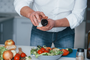 Putting some salt. Man in white shirt preparing food on the kitchen using vegetables