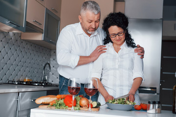 Happy together. Man and his wife in white shirt preparing food on the kitchen using vegetables