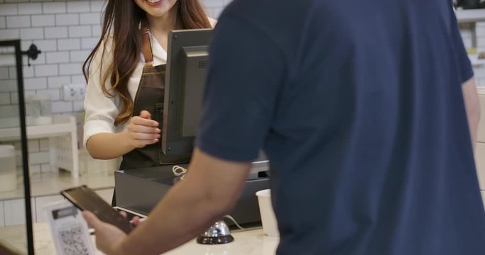 Beautiful asian woman serving a coffee to go to customer who uses smartphone to pay in coffee shop. QR code cashless payment concept.