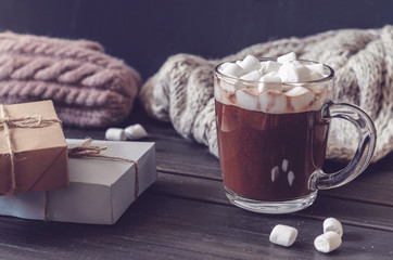 Hot cocoa with marshmallows in a glass cup surrounded by Christmas gifts and winter clothes,...