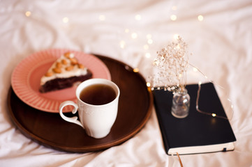 Cup of coffee, tasty cake with notebook and flowers in bed closeup. Good morning.