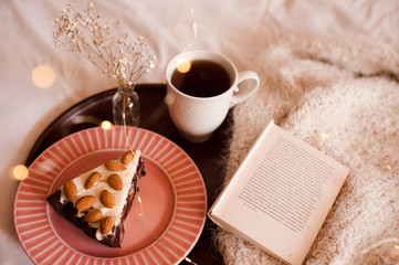 Cake with almond nuts on wooden tray with cup of tea and open book in bed. Good morning. Breakfast time.