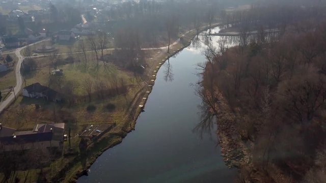Aerial: Flying towards river water mill at bank of beautiful green river. Filming beautiful river surroundings at spring sunset.