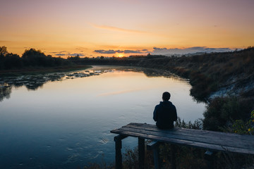 Naklejka na ściany i meble A man sitting alone on the old wooden bridge near the small countryside river and enjoying sunset on a calm evening. Natural scenery with Sura river in Penza region, Russia