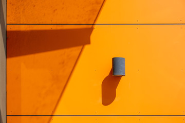 Exterior industrial wall mounted cylinder lamp on modern orange wall with strong shadows in bright...