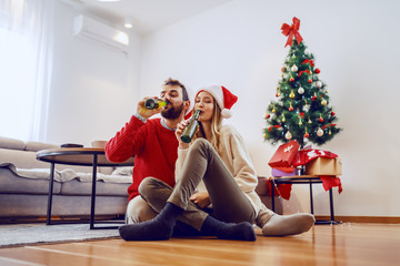 Adorable good-looking Caucasian couple with santa hats on heads sitting on floor in living room and drinking beer. In background is christmas tree with presents.