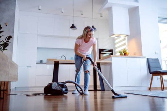 Smiling cheerful caucasian blonde housewife using vacuum cleaner to clean floor in living room. Home interior.