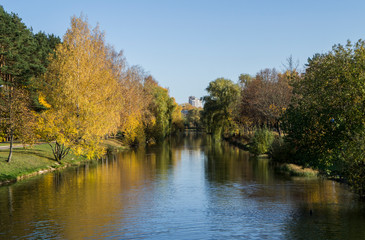 Autumn park with yellow leaves and trees with a river and reflection in the water and blue sky.
