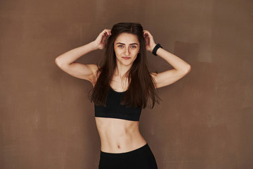 Fototapeta na wymiar Ready for the fitness exercises. Young beautiful woman standing in the studio against brown background