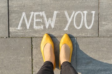 Text new you written on gray sidewalk with women legs, top view