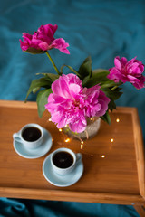 Beautiful pink peonies and two cups of coffee are standing on a wooden tray in bed.