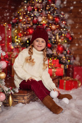 cheerful cute girl In a light knitted sweater and red hat sits on the steps of a wooden house in anticipation of the New Year holiday, in her hands she holds snowballs and has fun